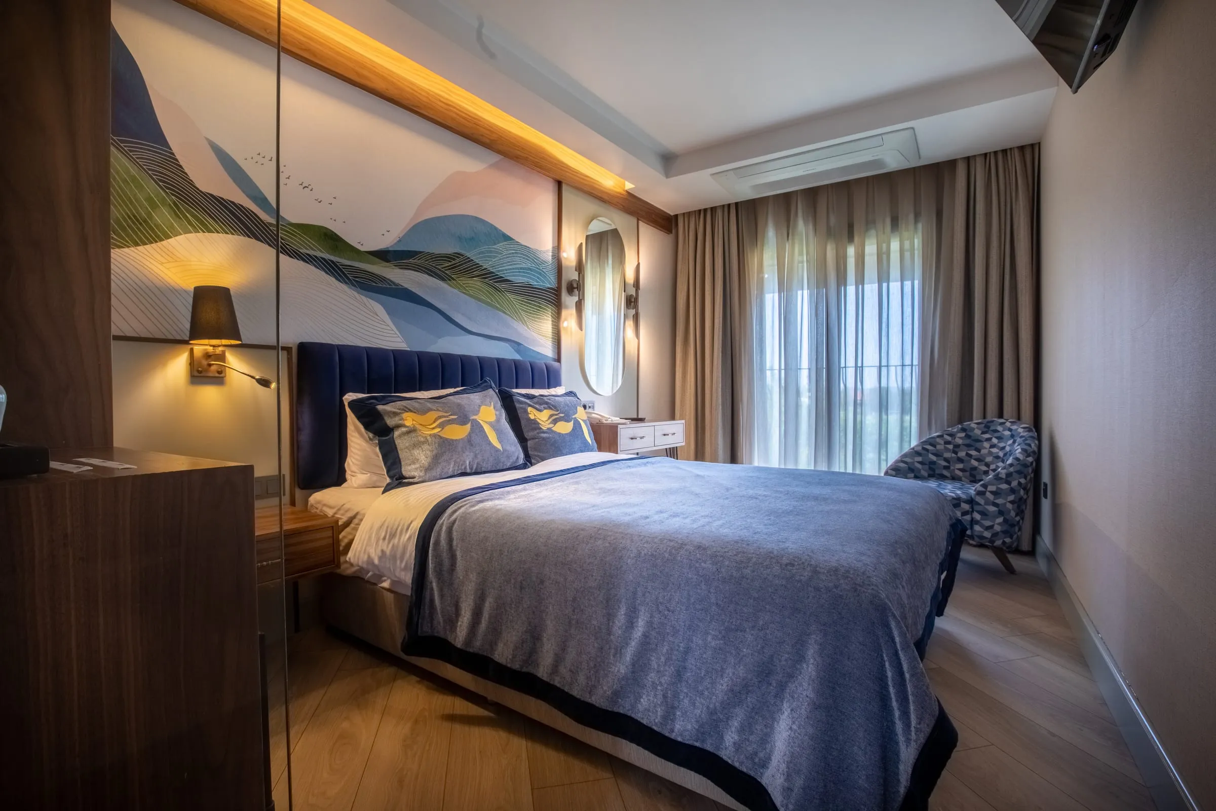 Petrol kadife başlıklı yatak, altın deniz kızı motifli yastıklar ve dalga muralı — otel odalarından biri